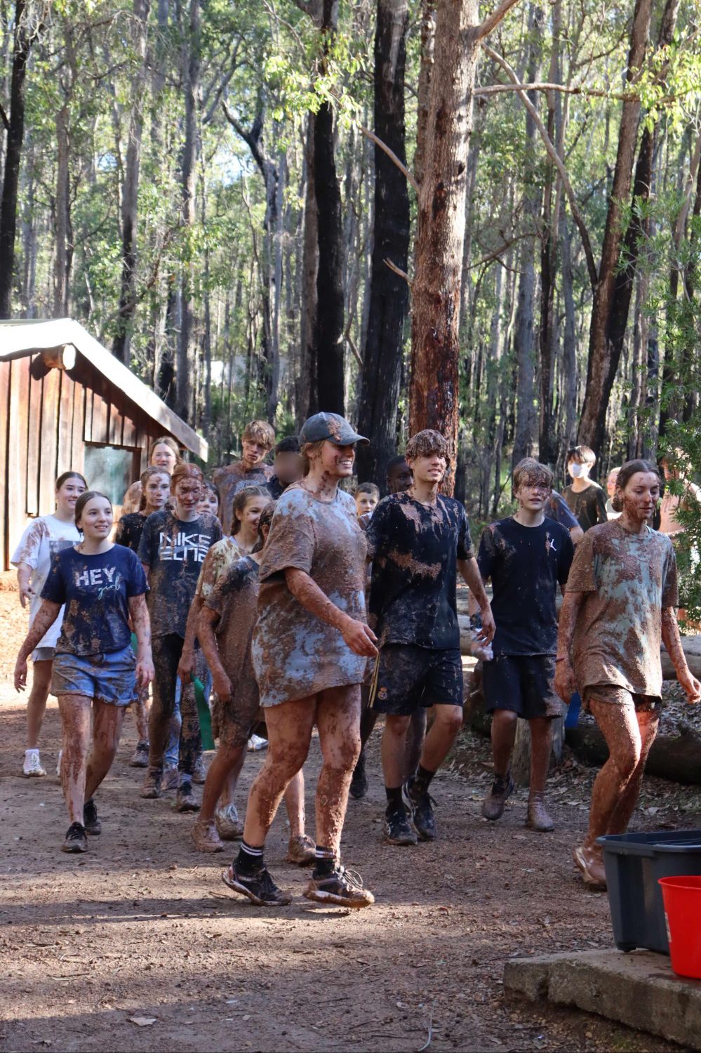 Teenagers covered in mud walk together smiling during an outdoor camp surrounded by tall trees.