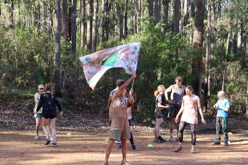 Campers celebrate outdoors after a muddy team game, with one person joyfully waving a large flag.