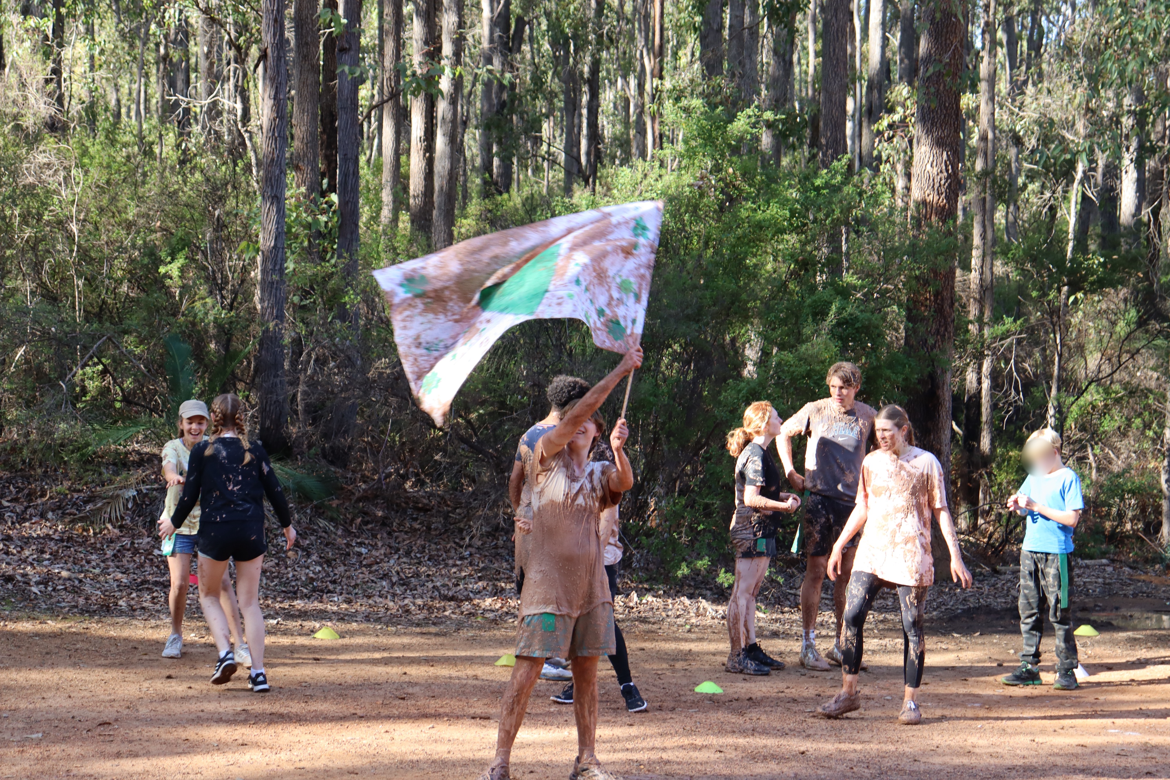 Campers celebrate outdoors after a muddy team game, with one person joyfully waving a large flag.