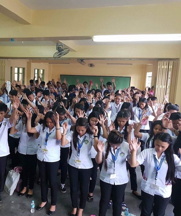 a group of teachers raise their hands and pray in a rustic classroom
