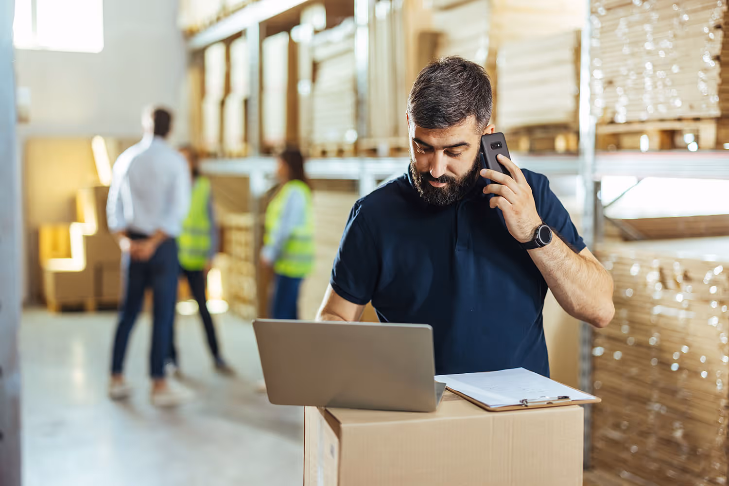Bearded man talking on phone while looking at laptop on a box in a warehouse with shelves of stacked goods and people in safety vests in the background.