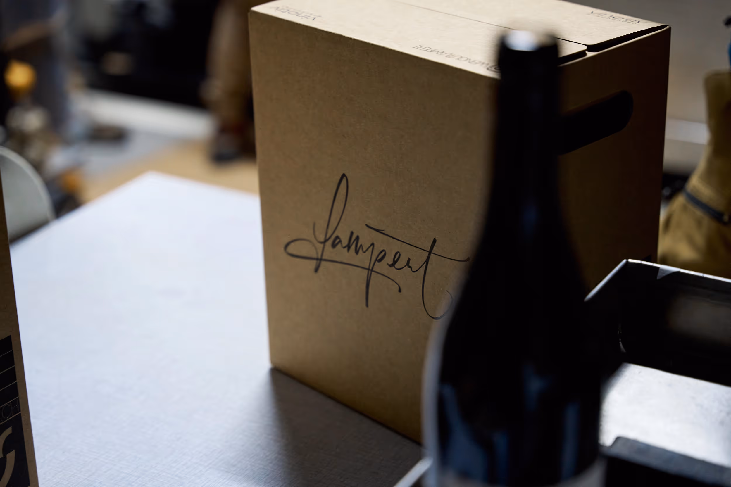 Flaschenkarton mit dem Lampert Logo steht auf einem Tisch und eine Weinflasche von Lampert davor