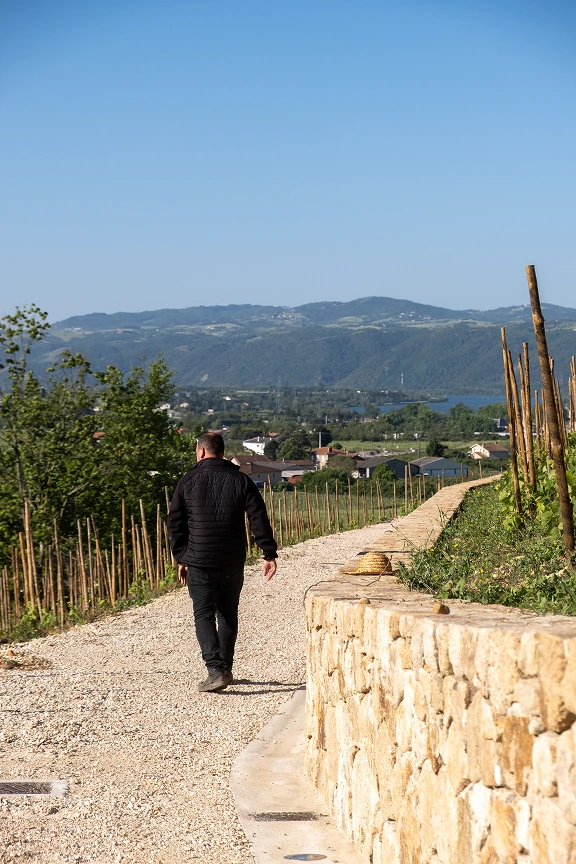 Yann Chave marche sur un chemin en gravier bordé de vignes, avec des montagnes et un village en arrière-plan sous un ciel bleu clair.