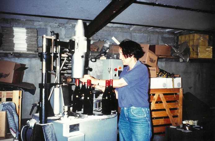 Une femme en chemise bleue embouteille du vin dans une cave avec des caisses en bois et des cartons en arrière-plan.