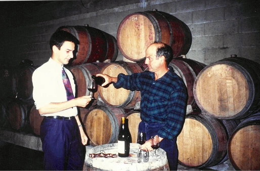 Deux hommes dans une cave à vin, l’un verse du vin d’une bouteille dans un verre tenu par l’autre, avec des tonneaux en bois empilés en arrière-plan.