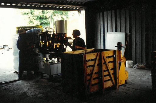 Deux personnes travaillent avec une machine dans un entrepôt sombre à côté d'une caisse en bois et d'un transpalette jaune.