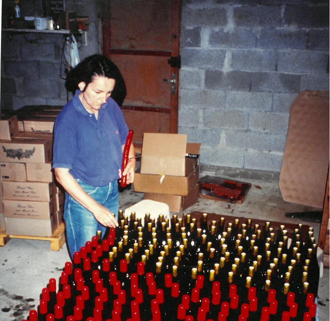 Femme rangeant des bouteilles de vin avec des capsules rouges et dorées dans un entrepôt en arrière-plan de murs en parpaings.