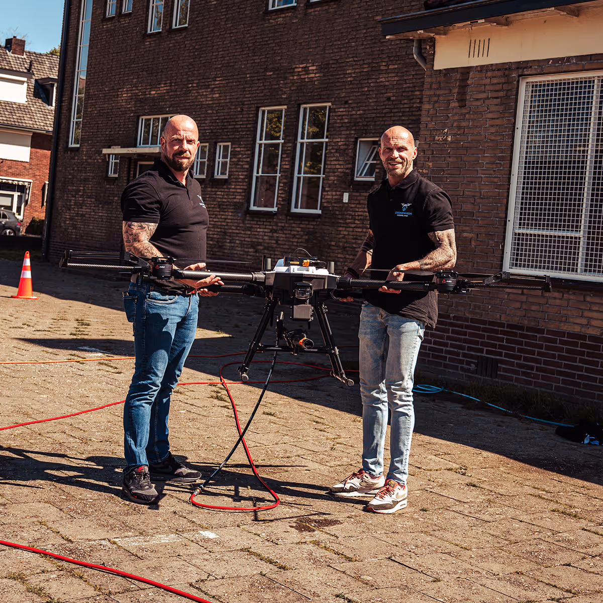 Twee mannen met tattoos houden een grote drone vast buiten naast een bakstenen gebouw.