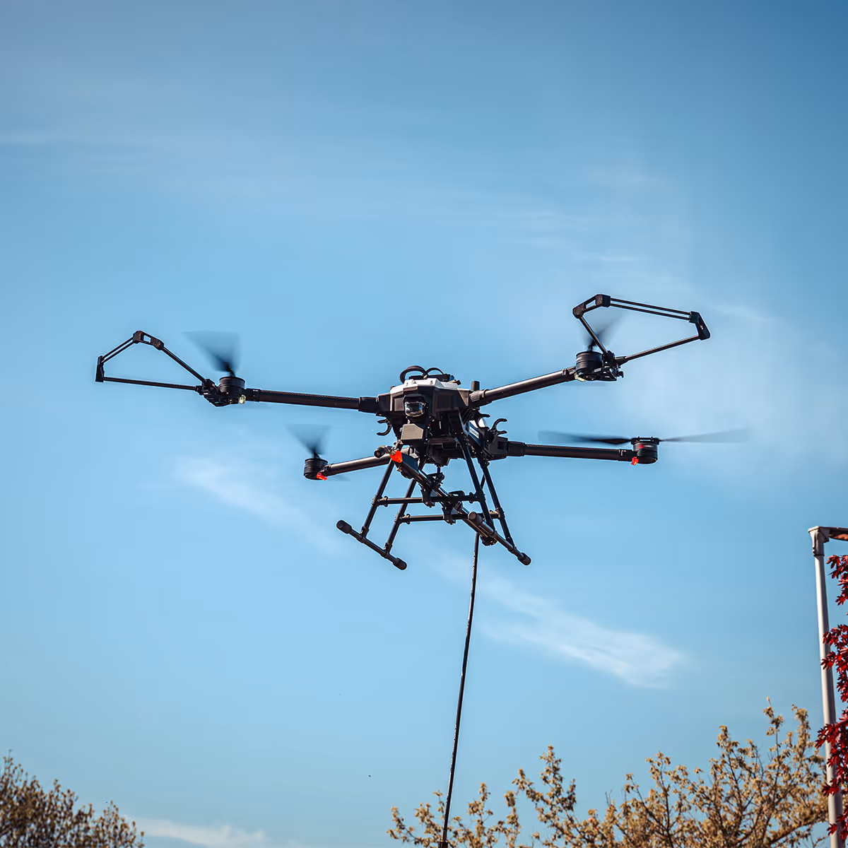 Zwarte drone met vier rotoren vliegt in de lucht boven bomen onder een blauwe lucht.