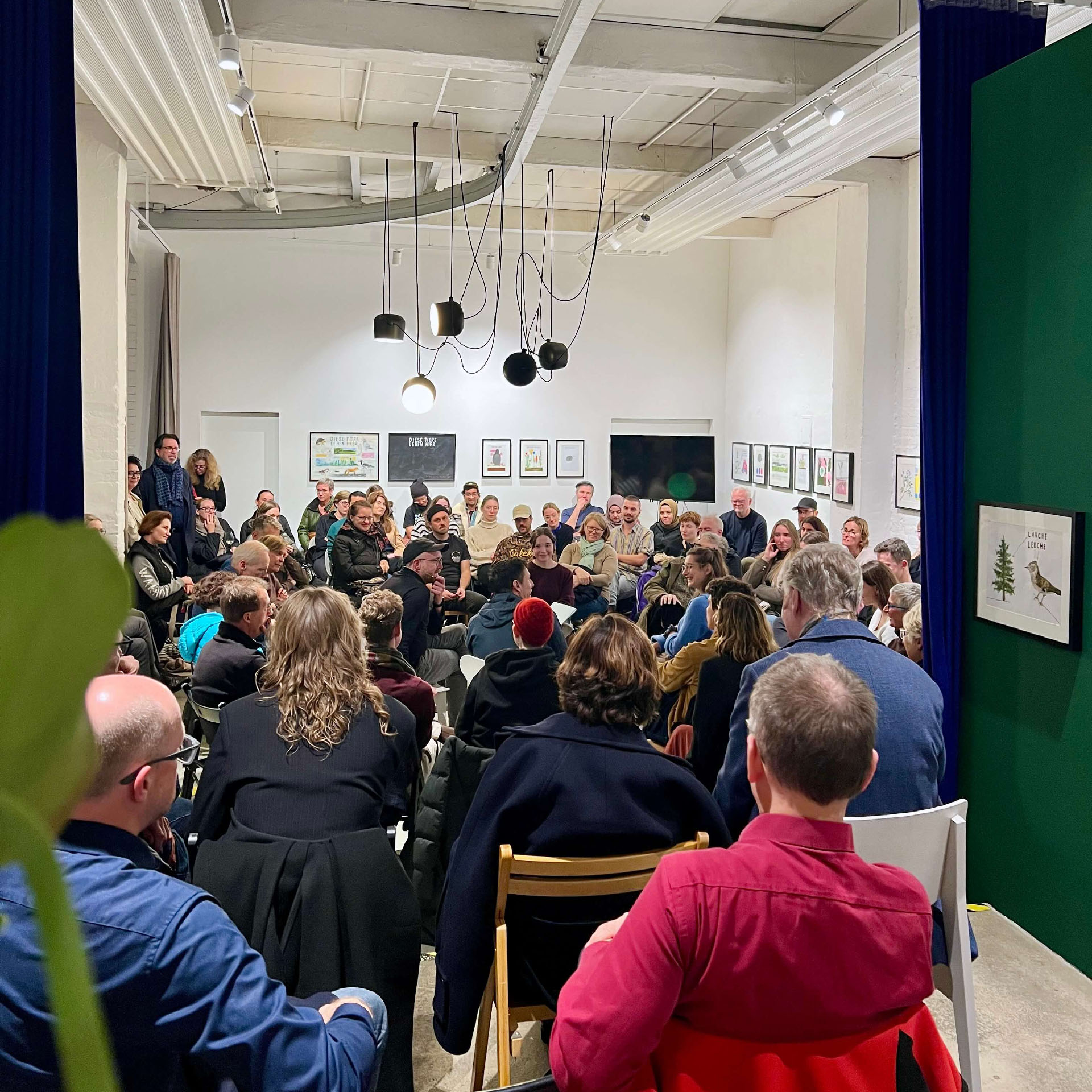 Blick von hinten in den voll besetzten Veranstaltungsraum bei GE59 und HENKELHIEDL. Das Publikum sitzt in einem dichten Stuhlkreis und blickt zur Mitte, im Vordergrund ist unscharf eine Pflanze zu sehen.