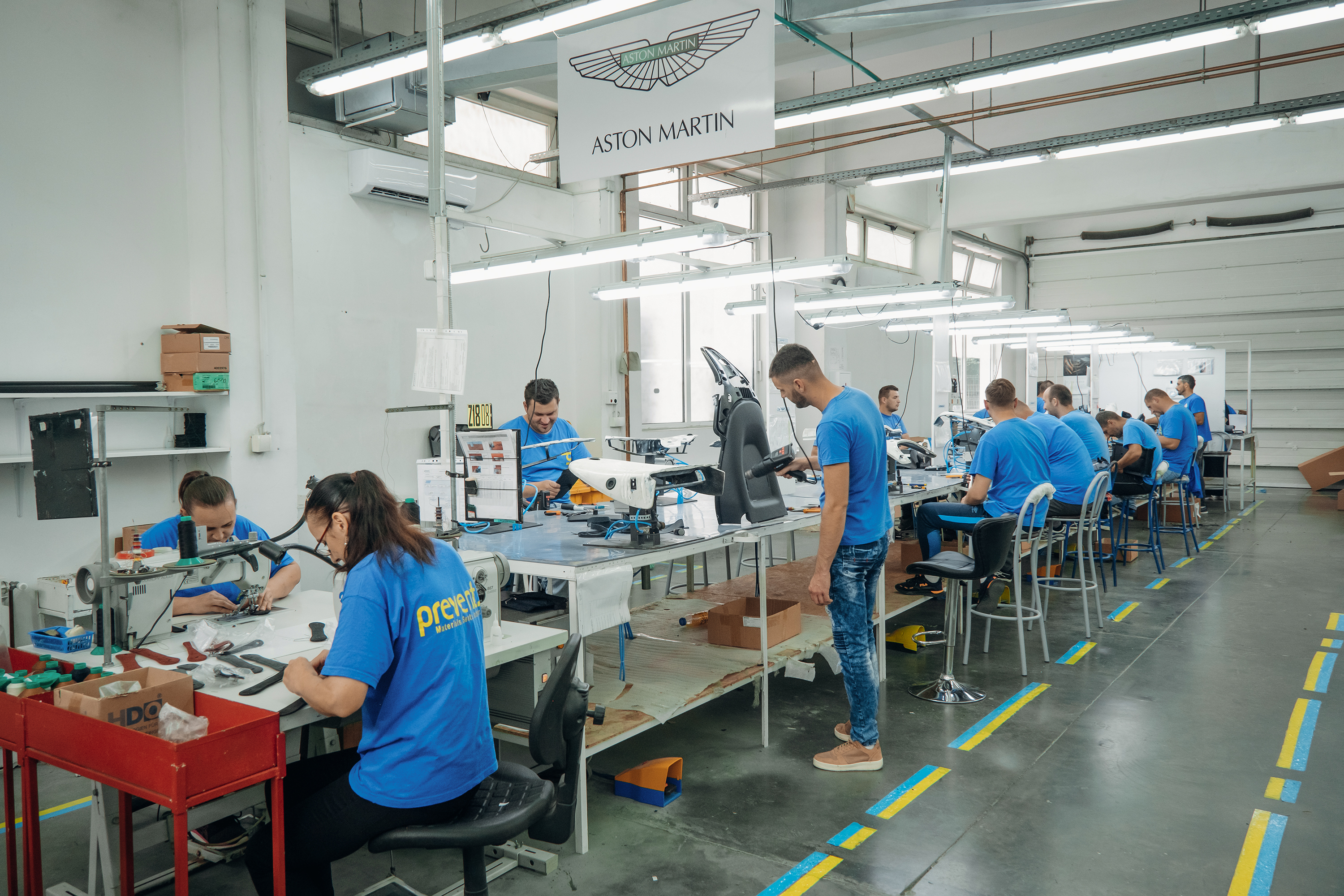 Workers in blue Prevent-branded shirts assemble automotive components at individual workstations in a clean, well-lit factory. A sign above reads “Aston Martin,” highlighting the brand partnership. The scene conveys precision and teamwork in a modern manufacturing environment.