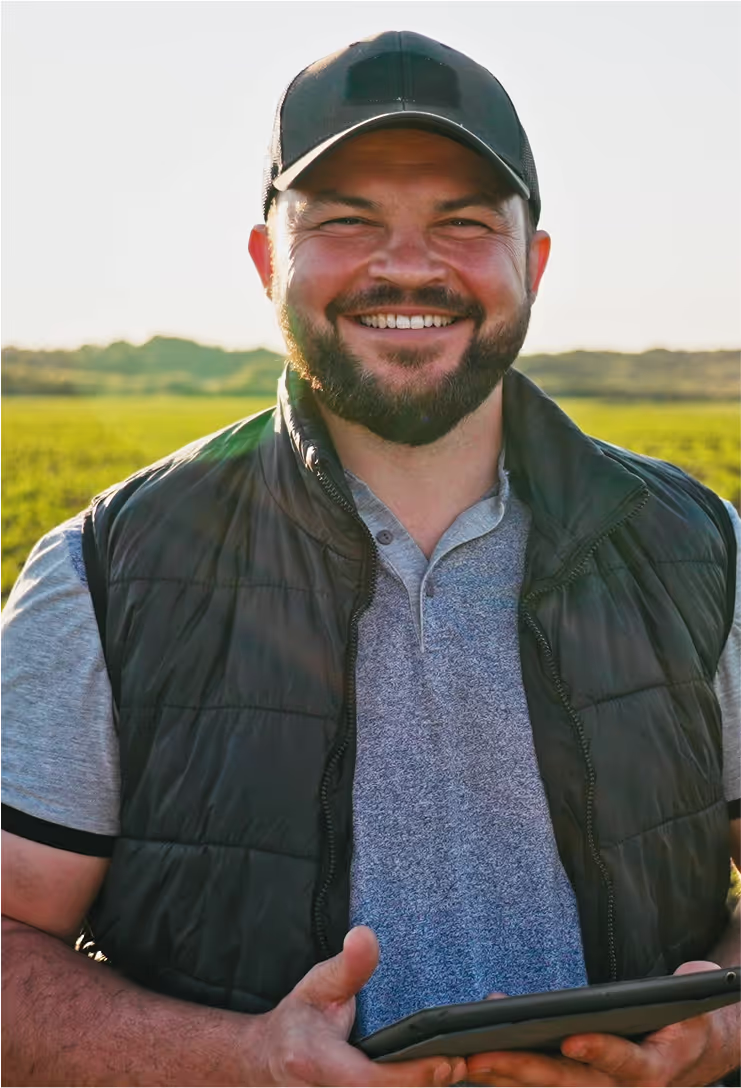 A cheerful bearded farmer wearing a cap and vest holds a tablet while standing in a green agricultural field