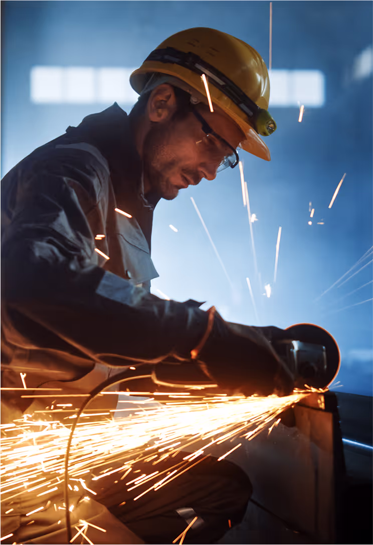 A worker in safety gear, including a yellow hard hat and goggles, uses a grinder, creating a shower of sparks in a dimly lit workshop