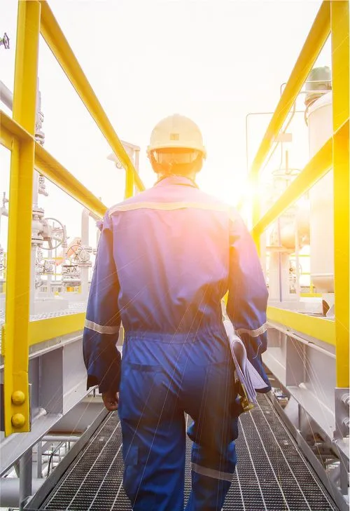 A worker in a blue jumpsuit and yellow hard hat walks along a high metal scaffold at an industrial site, bathed in bright sunlight
