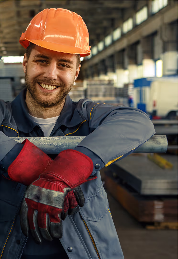 A smiling male factory worker in a hard hat and gloves leans on a metal pipe inside a manufacturing facility