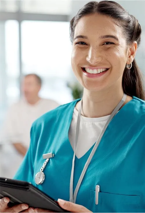 A smiling nurse in teal scrubs holds a tablet in a bright medical setting, wearing a stethoscope and ID badge