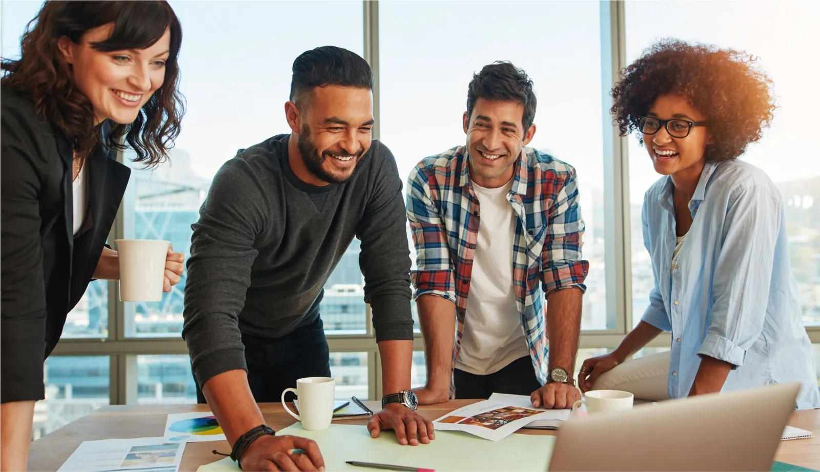 A diverse group of four professionals, two men and two women, are gathered around a table in a modern office, smiling and engaged in a discussion with documents, a laptop, and coffee cups in front of them.