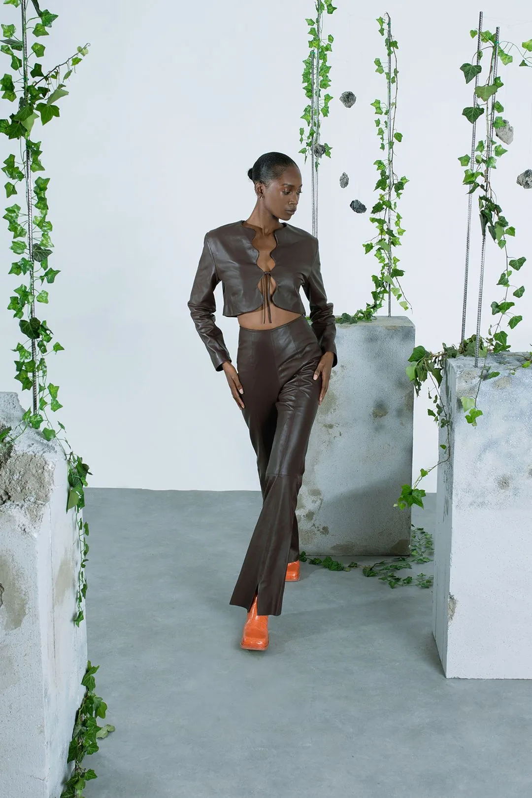 Fashion model wearing a dark brown leather crop jacket and flared trousers in a studio setting, showcasing high-end designer apparel by Prevent.