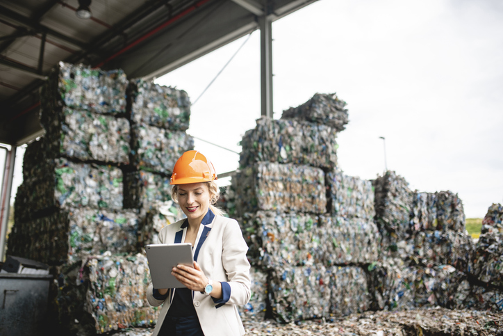 Female resource recovery specialist wearing an orange hard hat, using a digital tablet while standing outdoors near industrial recycling equipment.