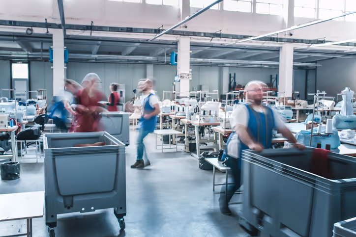 Workers pushing carts near sewing machines in a busy textile factory filled with fabric, garments, and industrial equipment.