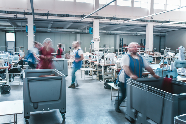 Workers pushing carts near sewing machines in a busy textile factory filled with fabric, garments, and industrial equipment.