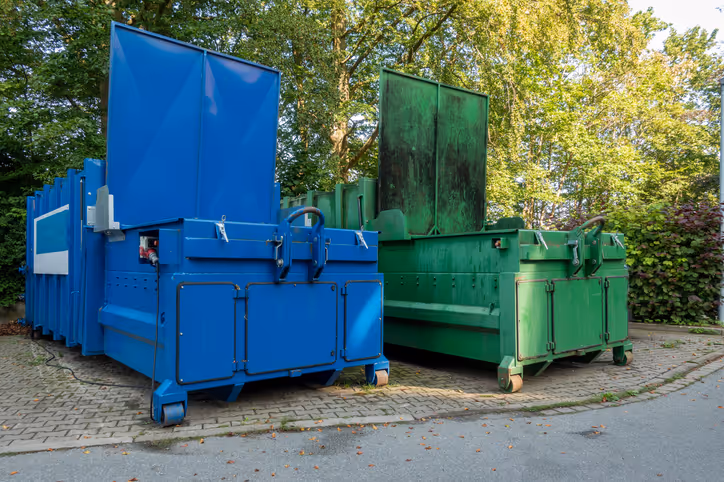 Two large garbage compactors positioned side by side on a brick pad with trees in the background.
