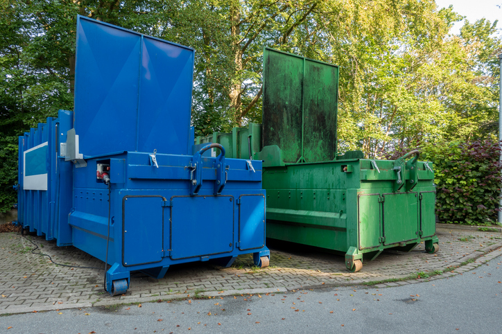Two large garbage compactors positioned side by side on a brick pad with trees in the background.