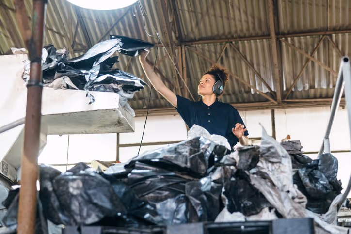 Female recycling facility worker wearing hearing protection reaching into a pile of black plastic materials under a corrugated metal roof.