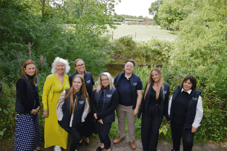 Group of eight women, who all work for Angel Accounting, standing outdoors on a path surrounded by green trees and bushes with a farm visible in the background.