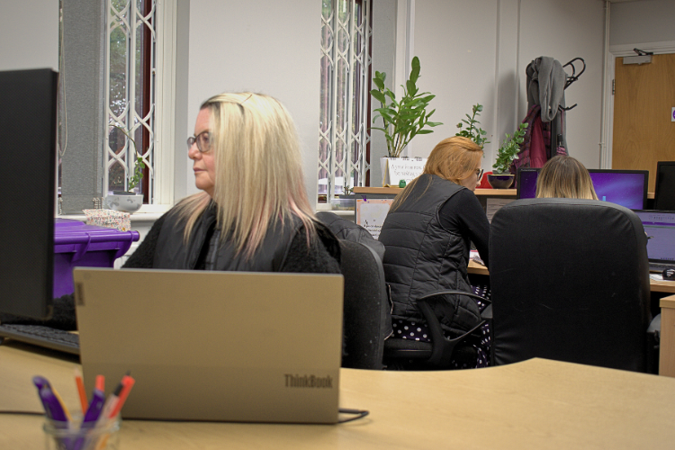Three women, who all work for Angel Accounting, working at desks with laptops and computers in an office space with plants and coats hung near a door.