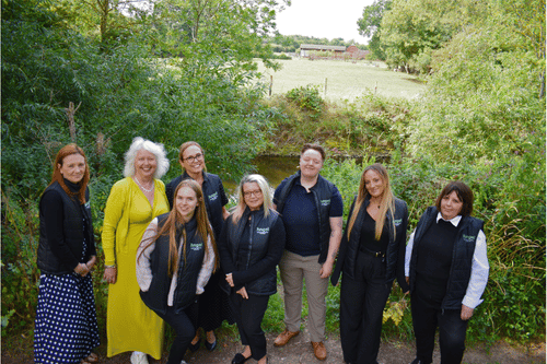 Group of eight women, who all work for Angel Accounting in Alcester, standing outdoors on a path surrounded by green trees and bushes with a farm visible in the background.