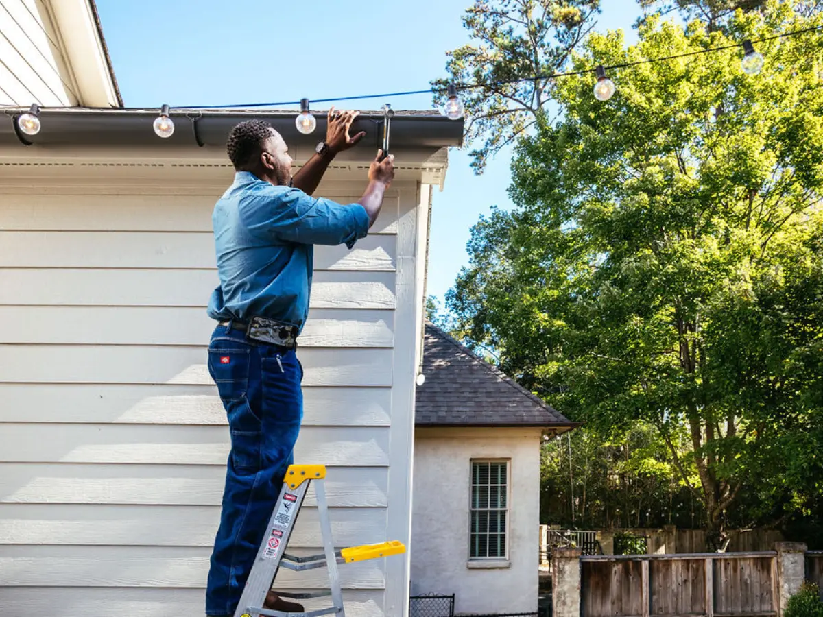  A man on a ladder adjusts a string of lights, demonstrating a home improvement task in a well-lit environment, image