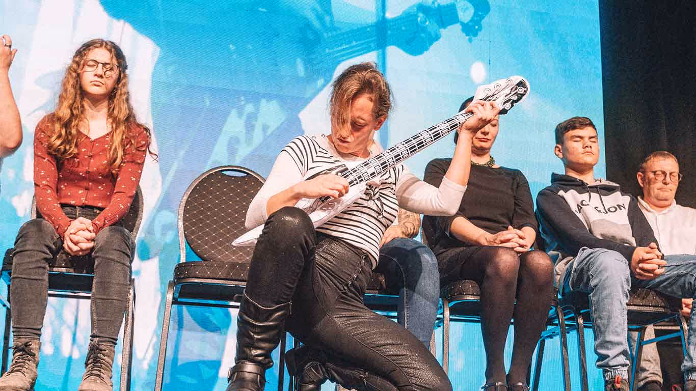 Person im gestreiften Hemd spielt auf einer aufblasbaren Gitarre vor einer Gruppe meditierender, Augen geschlossener Menschen auf Stühlen.