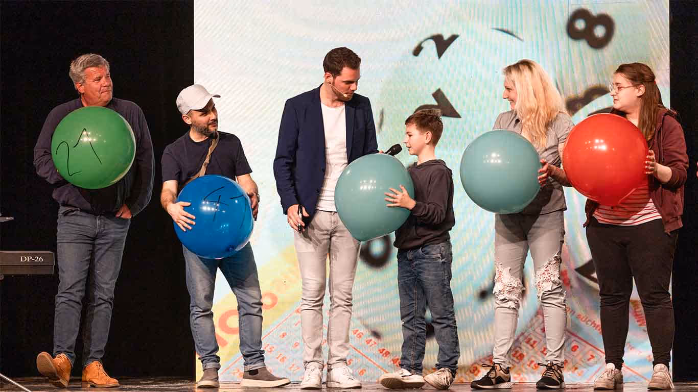 Sechs Personen unterschiedlicher Altersgruppen stehen auf einer Bühne, jeder hält einen großen Ballon in verschiedenen Farben, ein Junge spricht in ein Mikrofon.