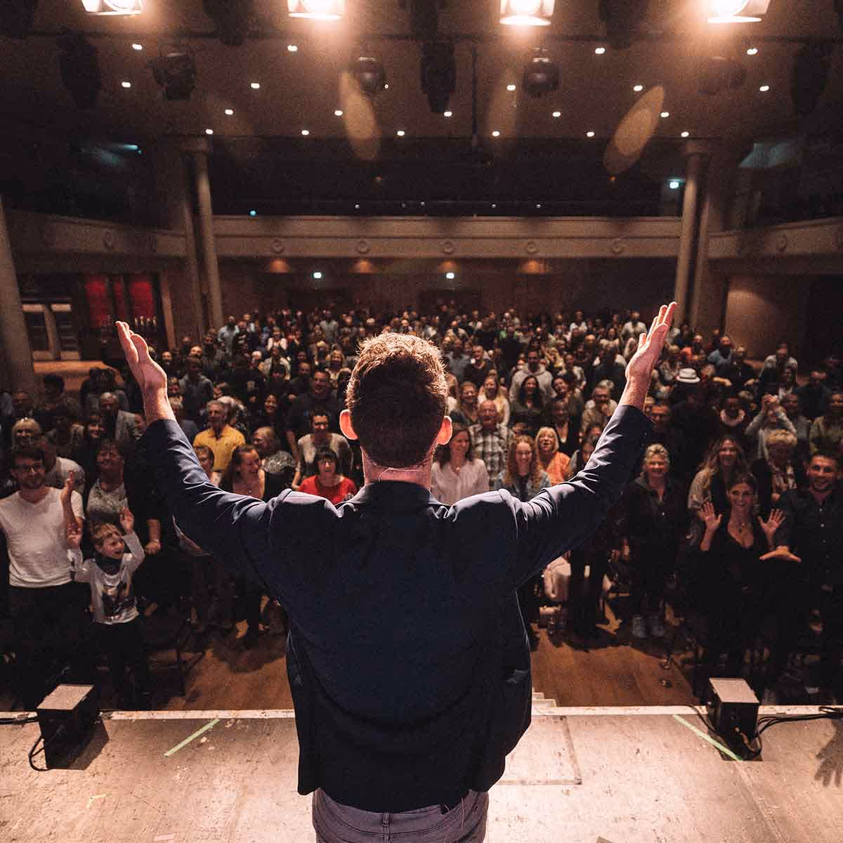 Ein Mann steht auf einer Bühne mit erhobenen Armen vor einem applaudierenden Publikum in einem Theater.
