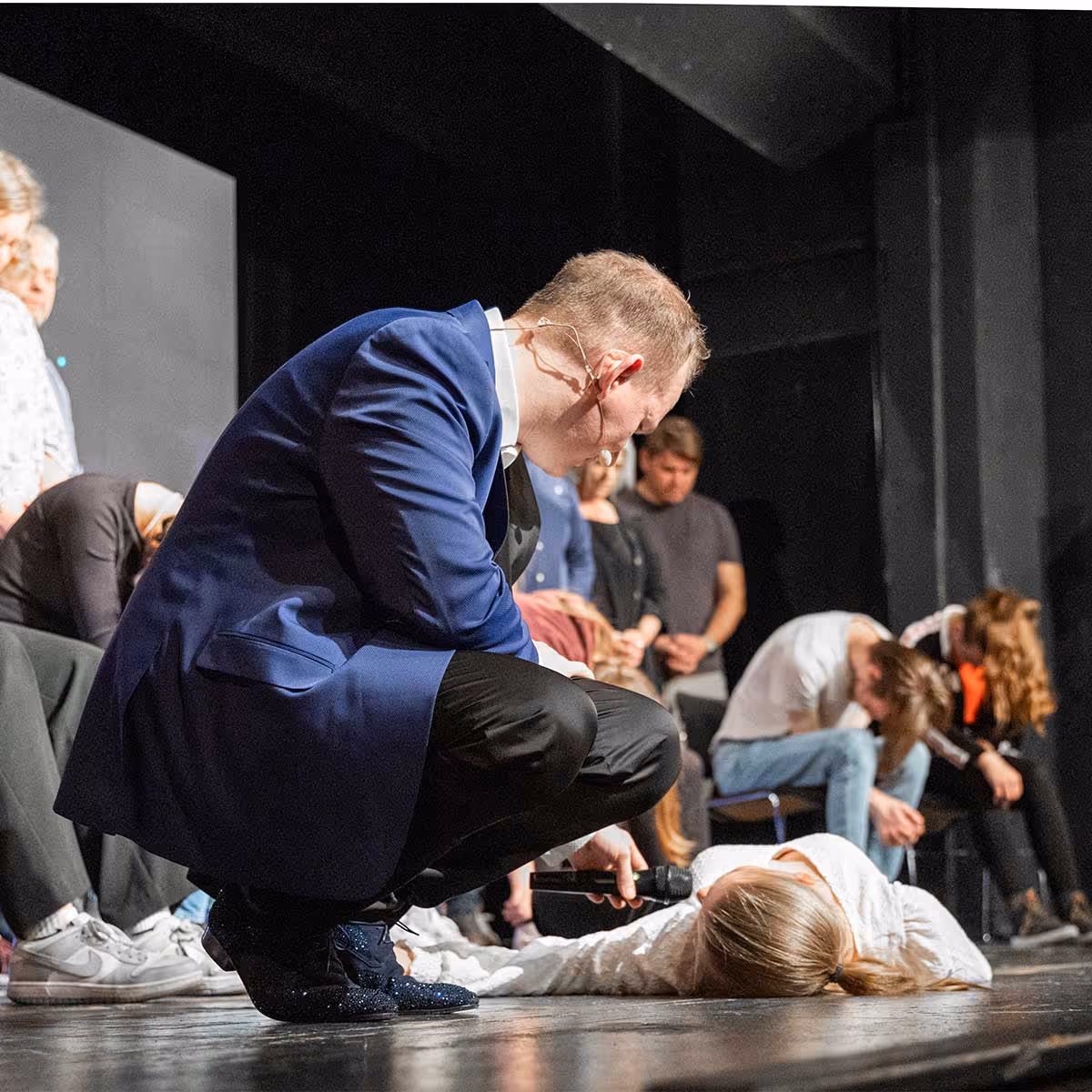 Ein Mann in blauem Anzug kniet und hält ein Mikrofon zu einer Person, die auf dem Bühnenboden liegt, während andere Menschen im Hintergrund sitzen und den Kopf gesenkt haben.