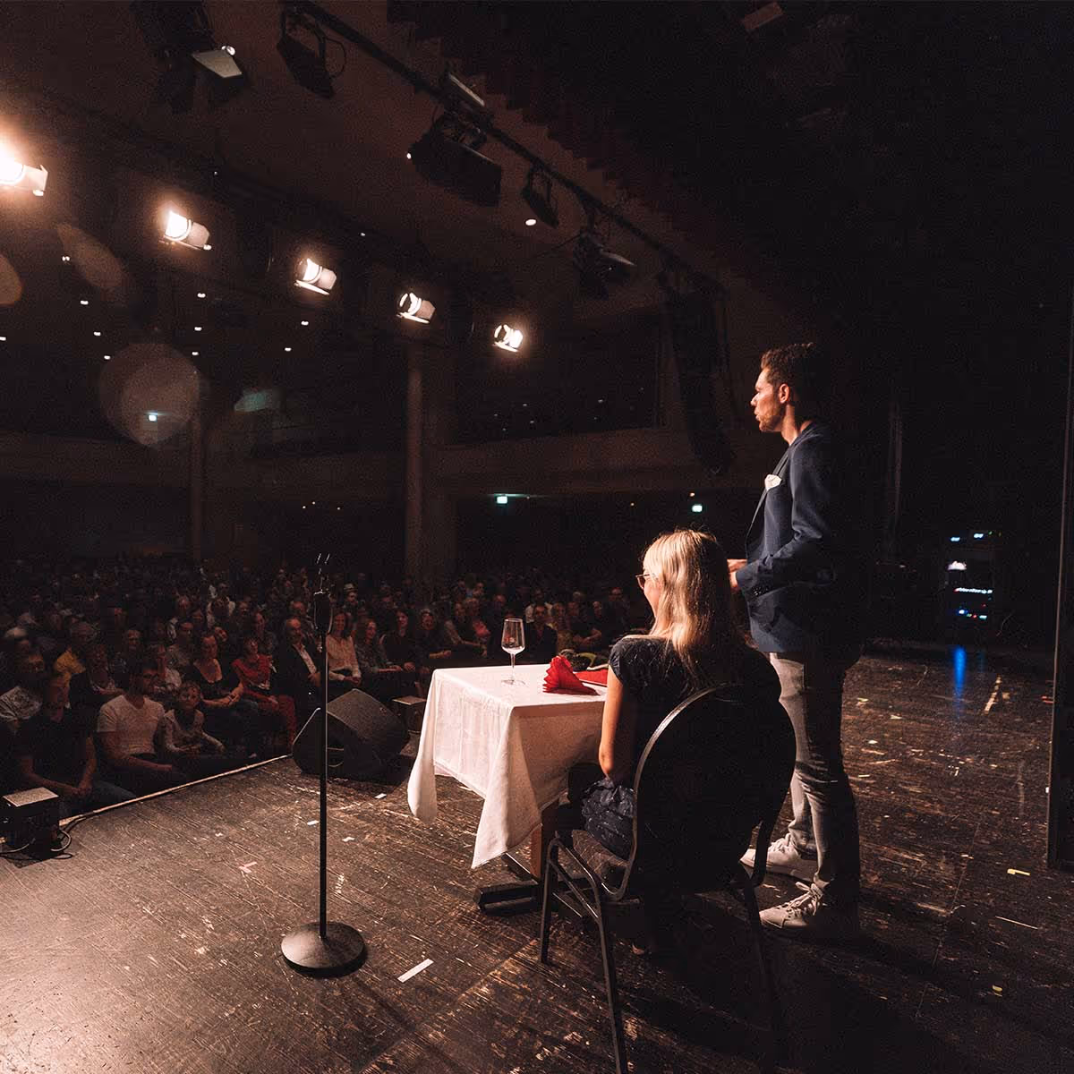 Eine Frau sitzt an einem Tisch mit weißer Tischdecke auf einer Bühne, während ein Mann neben ihr spricht und ein Publikum zuschaut.