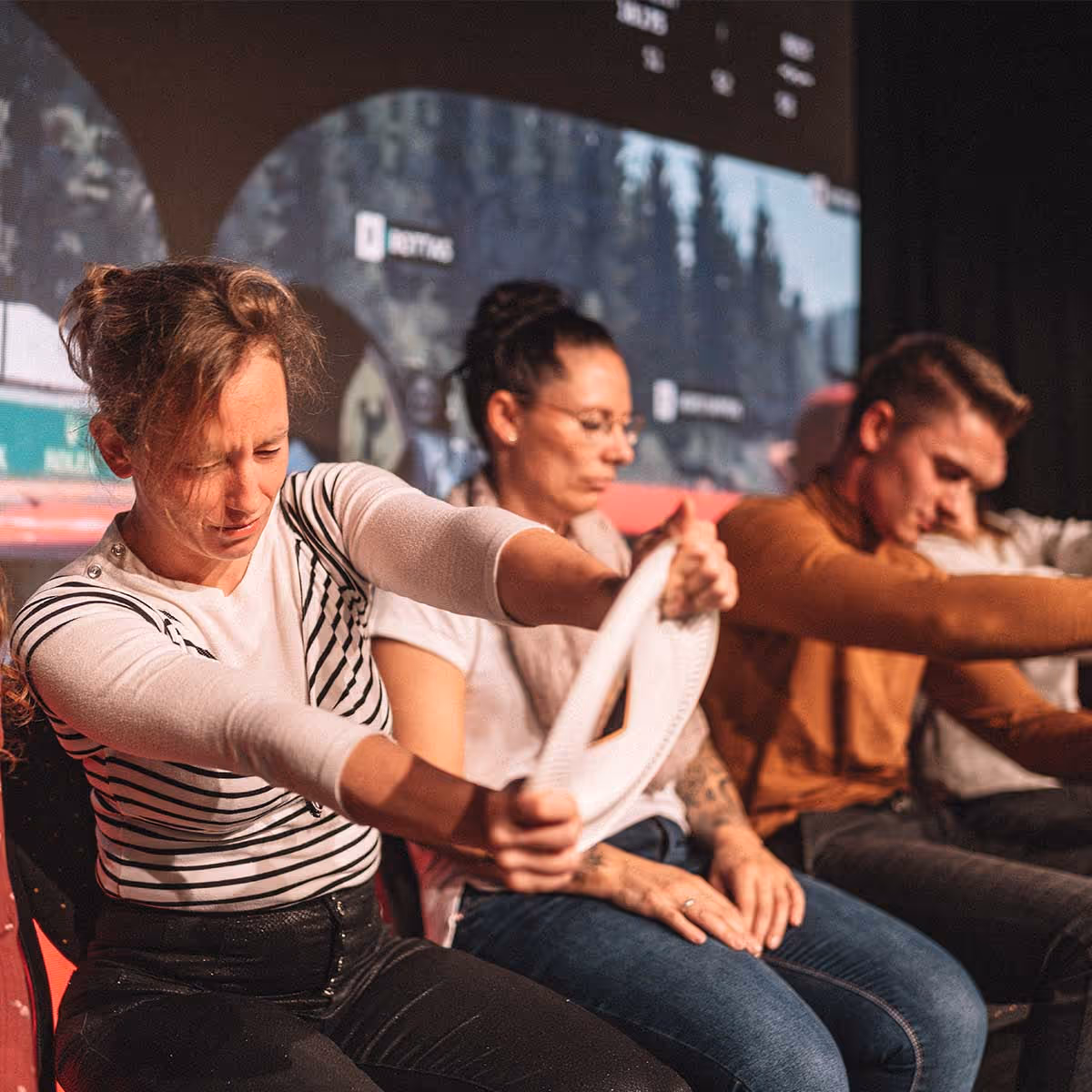 Eine Frau in einem gestreiften Shirt hält konzentriert ein weißes Tuch nach vorne, während andere Personen neben ihr sitzen und ebenfalls die Augen geschlossen haben.