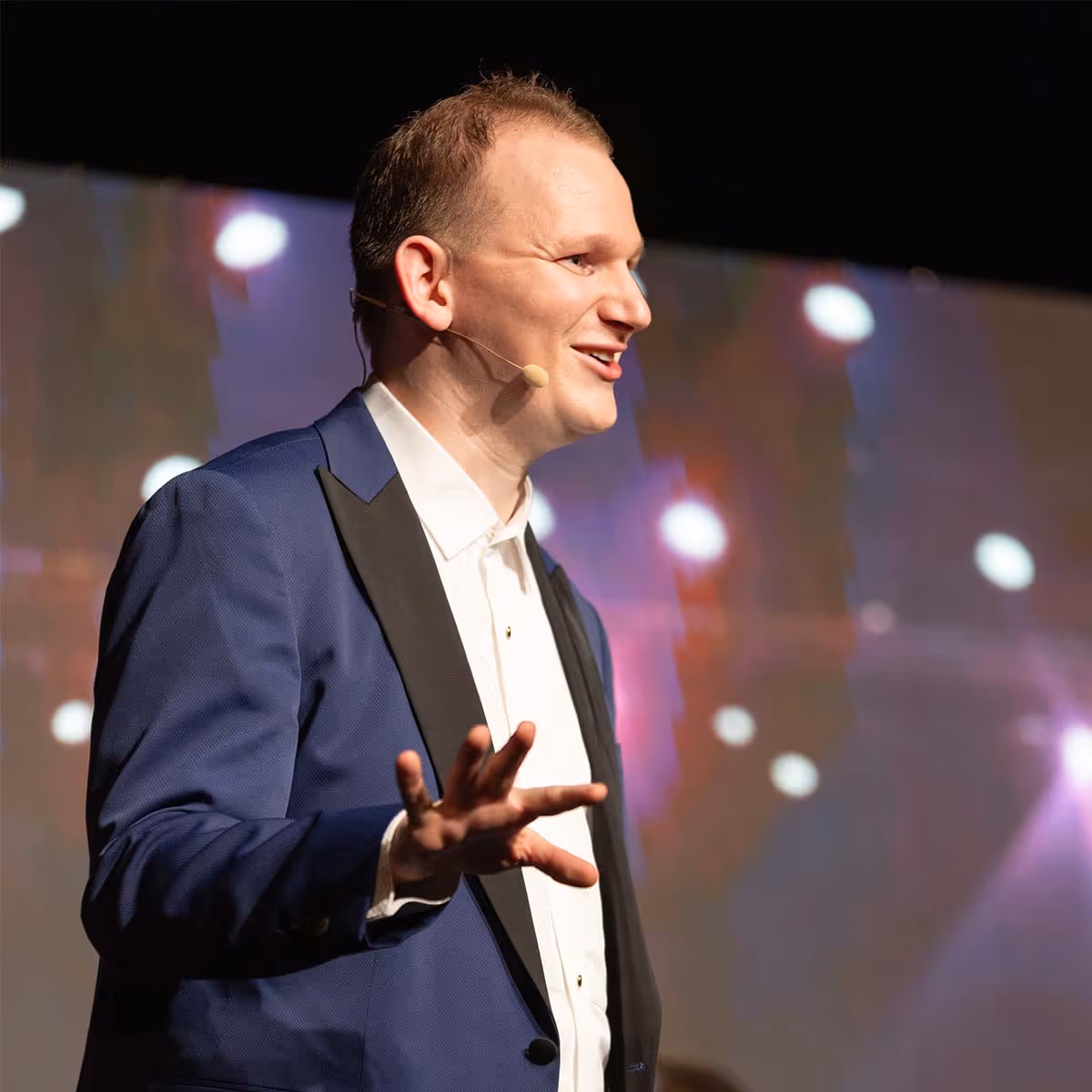 Mann im blauen Anzug mit Ansteckmikrofon spricht auf einer Bühne mit verschwommenen Lichtern im Hintergrund.