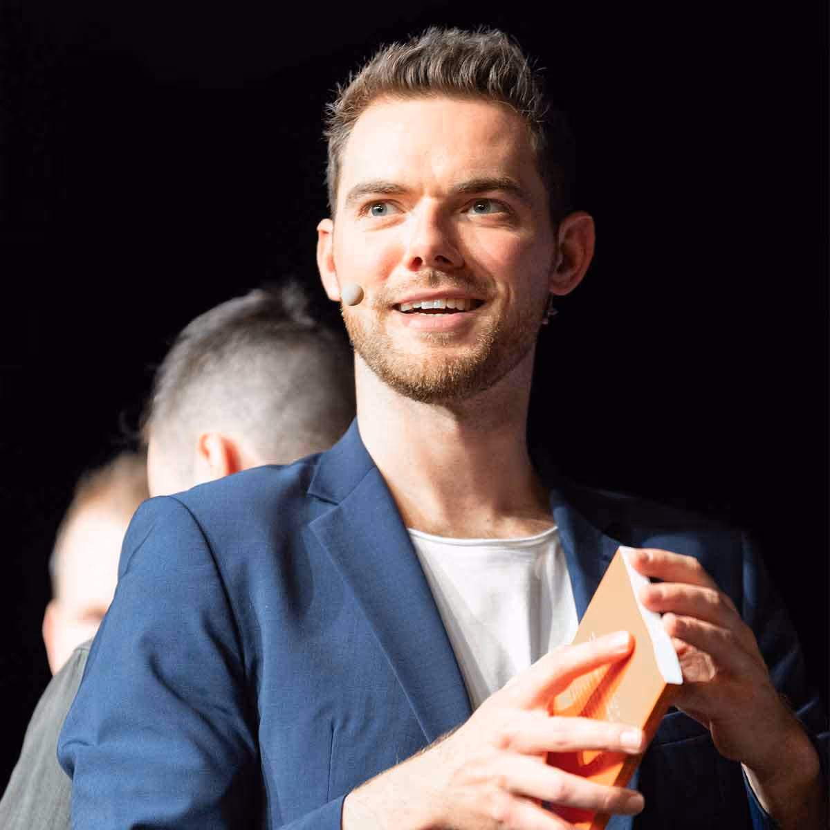 Junger Mann mit Mikrofon am Kopf hält ein orangenes Buch und lächelt.