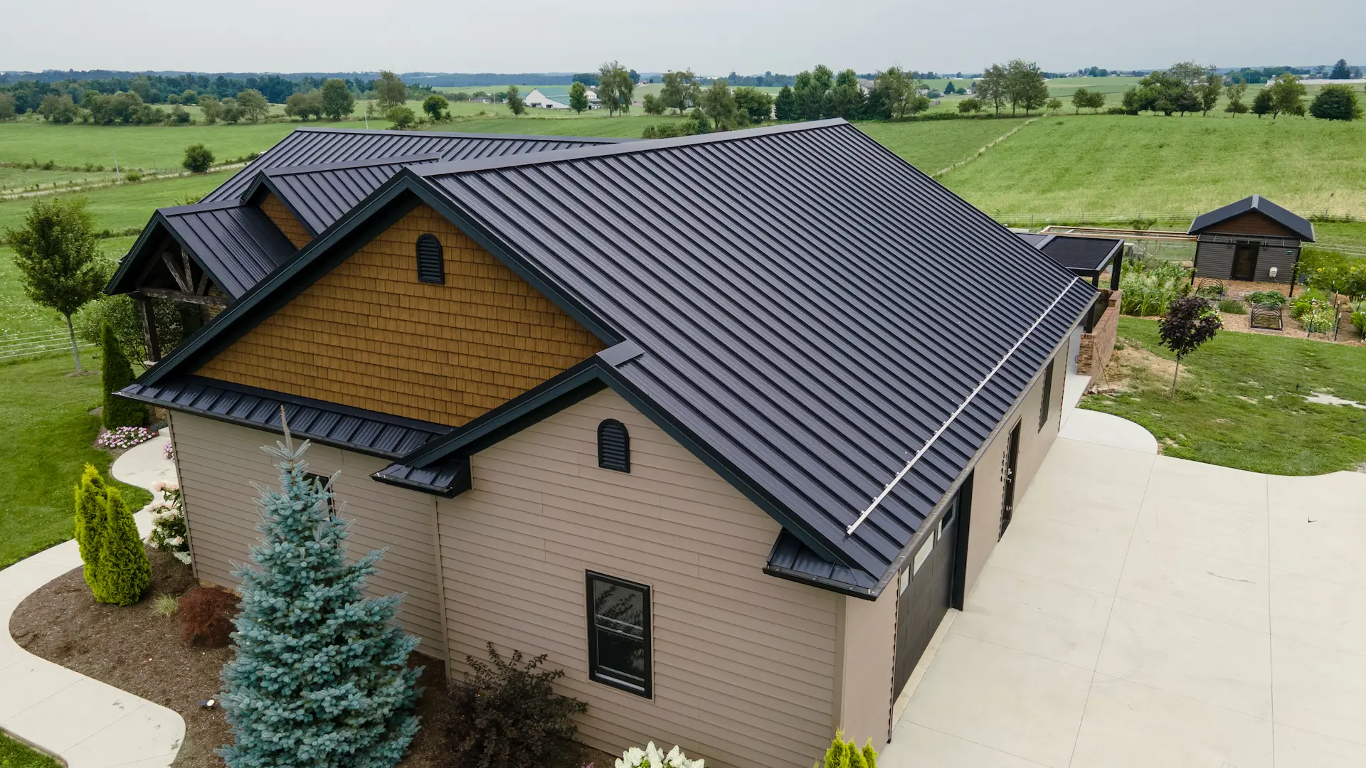 Aerial view of a custom home with a black standing seam metal roof, wood shake accents, and beige siding surrounded by green countryside.