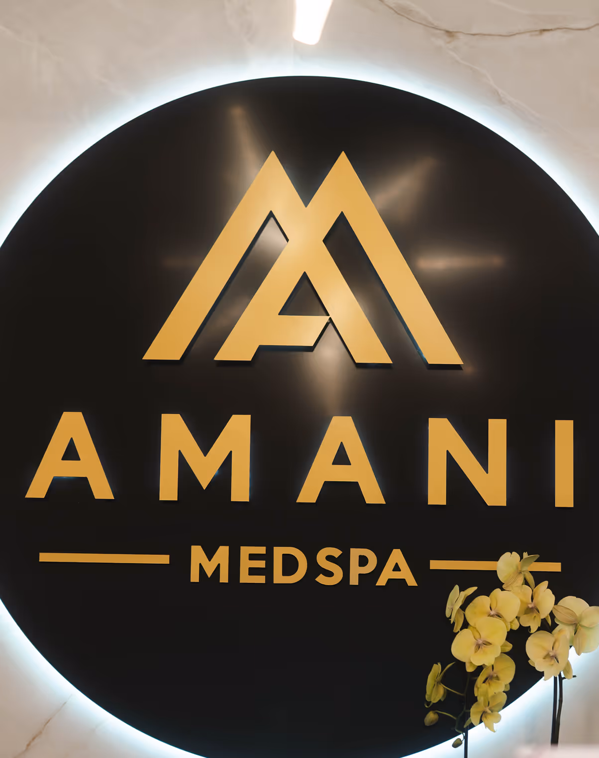 Black circular sign with gold letters spelling AMANI MEDSPA and a geometric double M logo above, with yellow orchids on the lower right.
