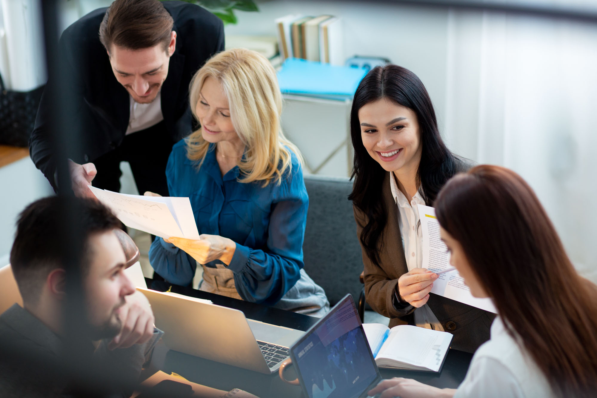 Five people smiling and discussing documents at a table with laptops.