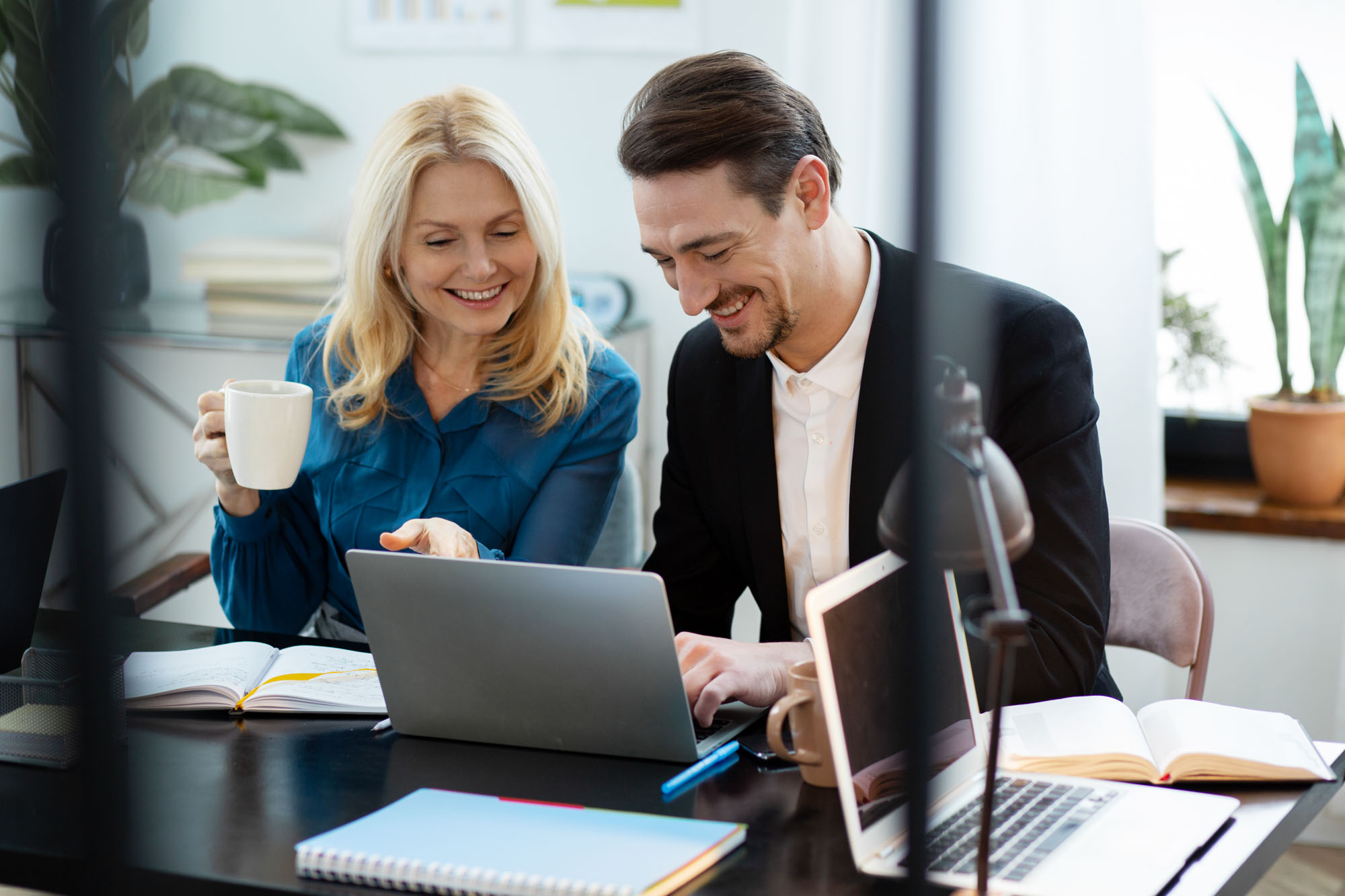 A woman and a man sit at a table, looking at a laptop and smiling.