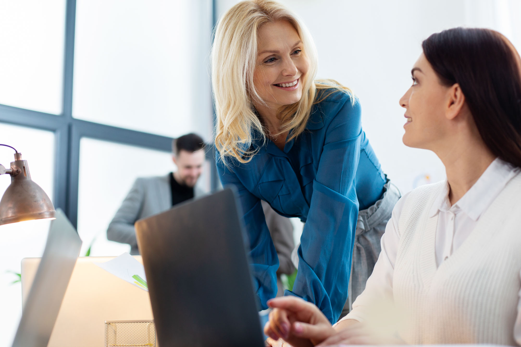 Two women in an office are talking and smiling at each other near a desk.