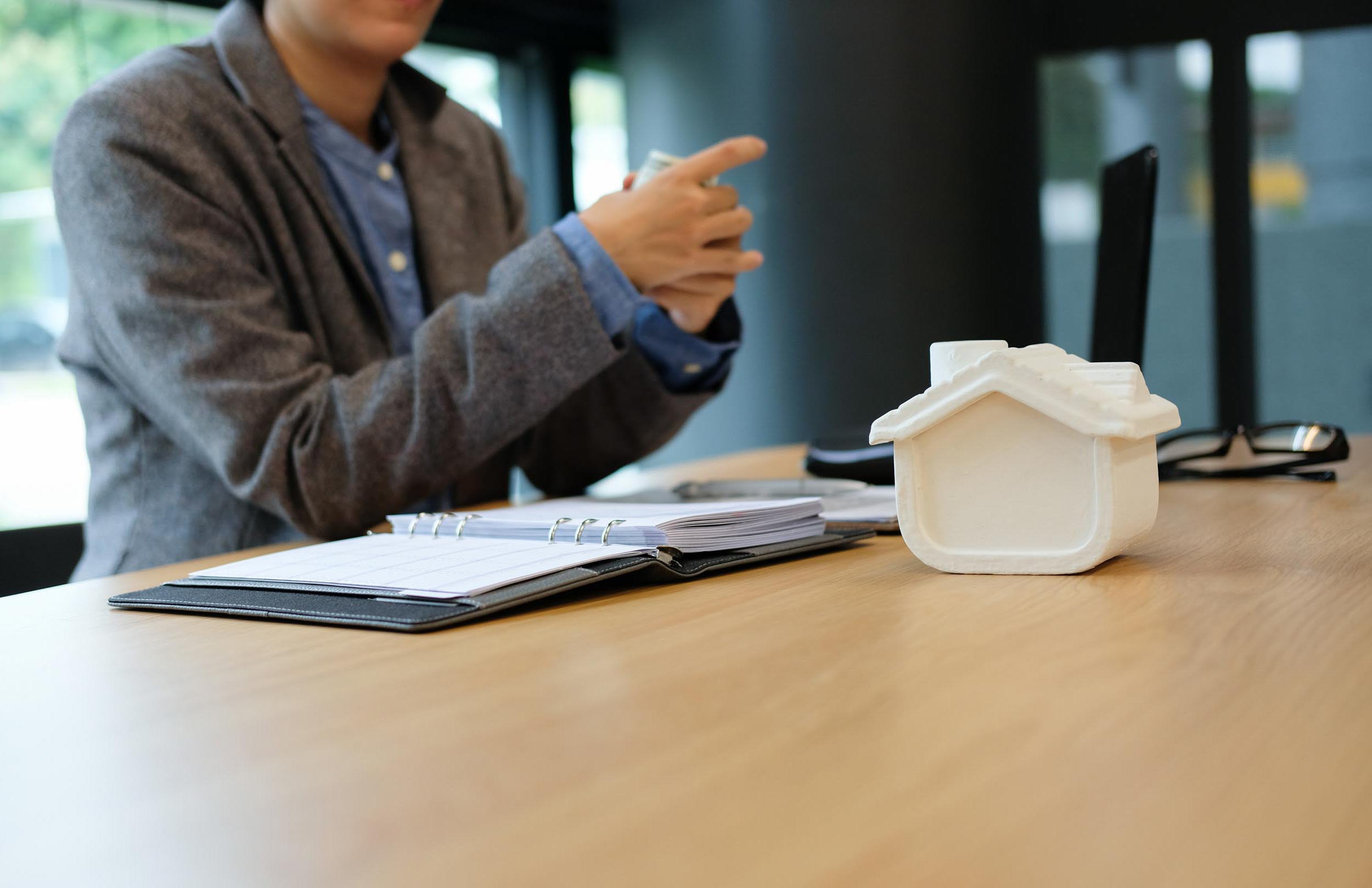 A person in a blazer sits at a desk with a notebook and small house model.