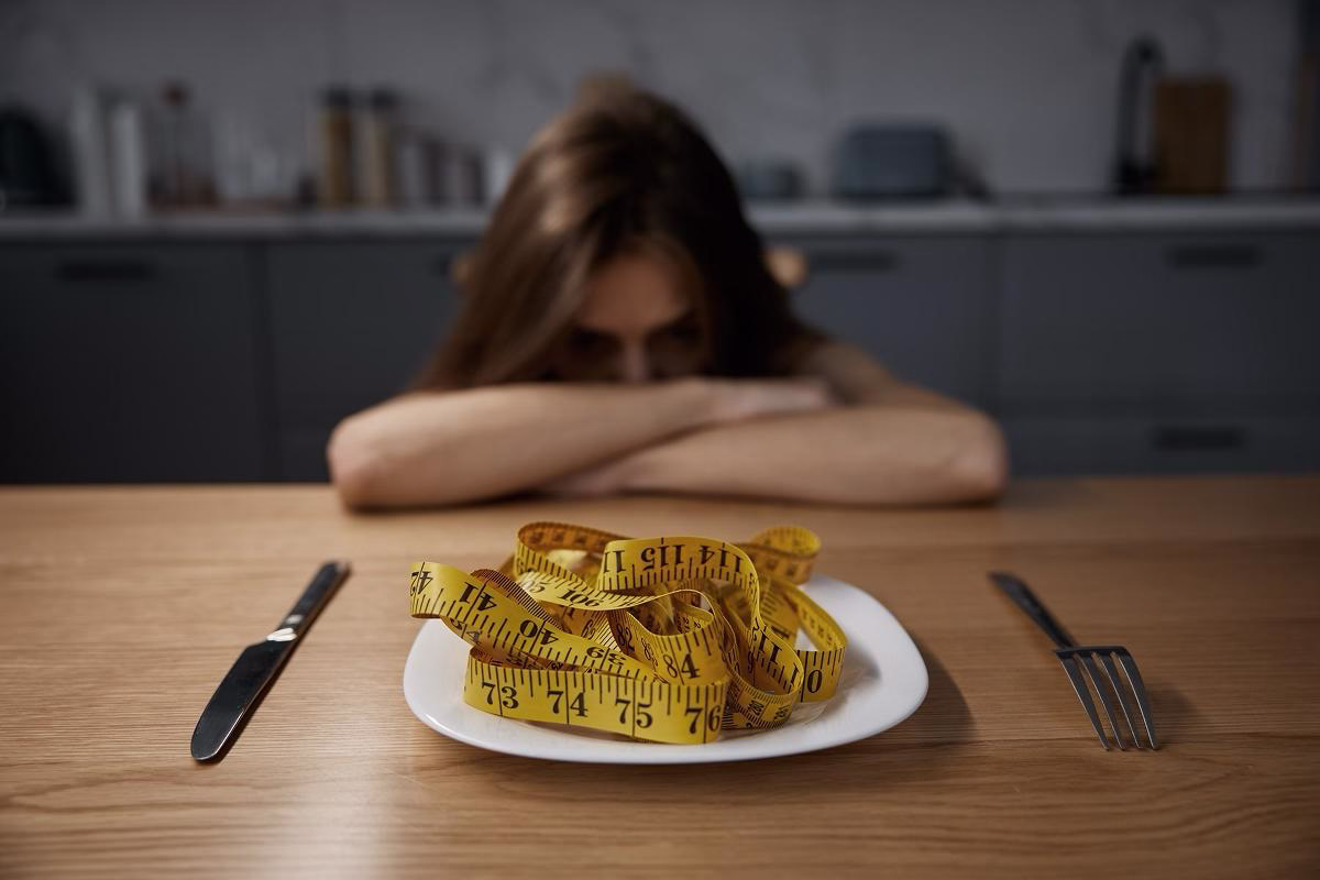 Femeie privește trist o farfurie cu un metru de croitorie, simbol al presiunii dietelor și al nemulțumirii față de corp.