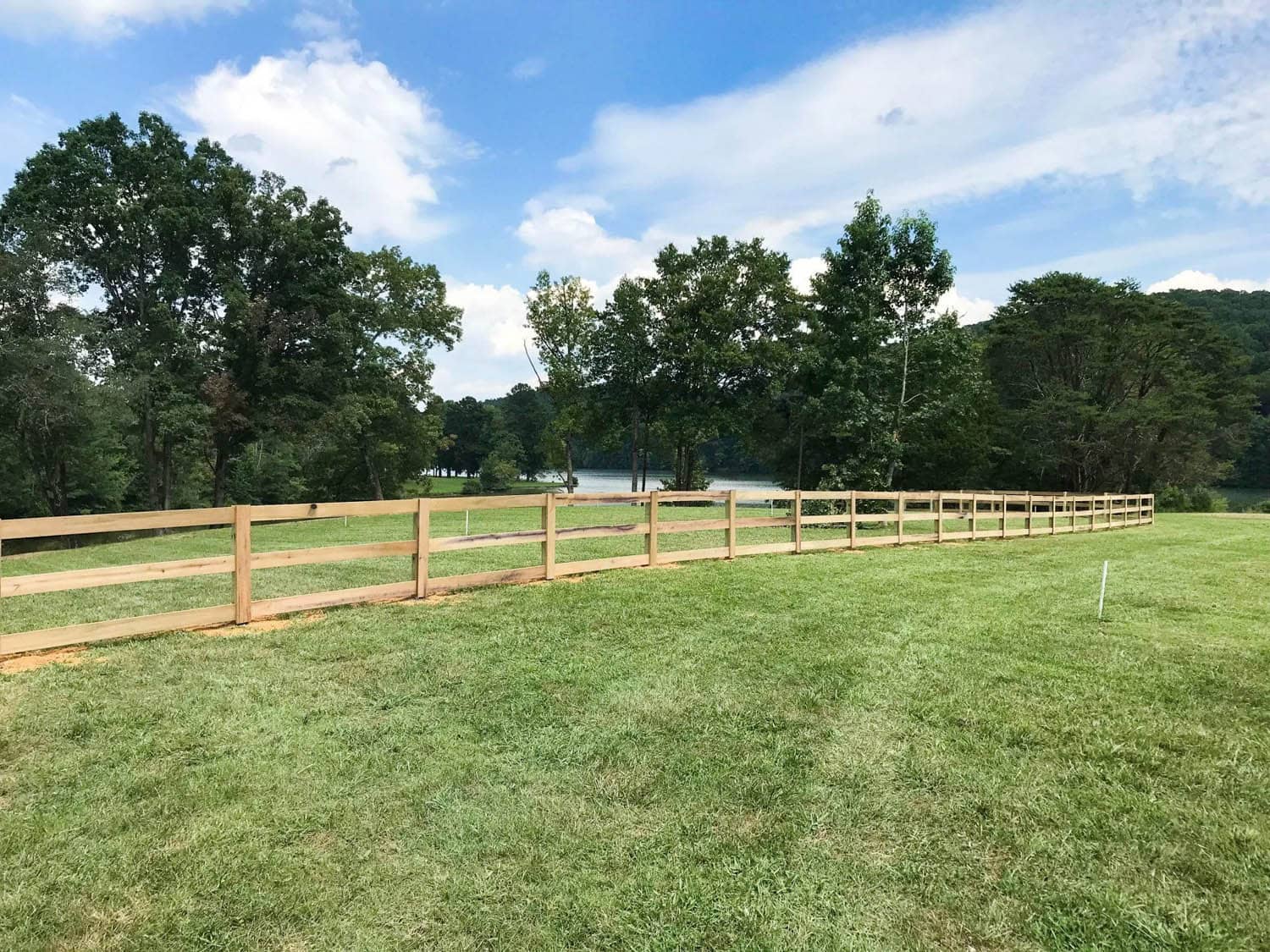 New three-rail wood fence running along a surveyed property line on an East Tennessee lakeside lot, with a white line stake in the grass, built by Loudon County Fence