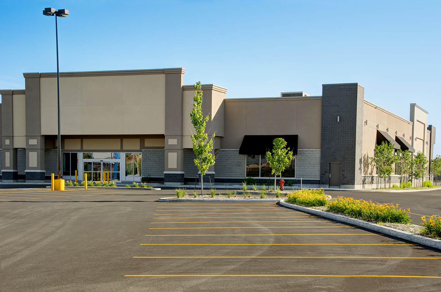A wide outdoor shot of a commercial building, with a mix of beige and grey colors, and a black awning. A large, empty asphalt parking lot with yellow lines and young trees is in the foreground under a clear blue sky.
