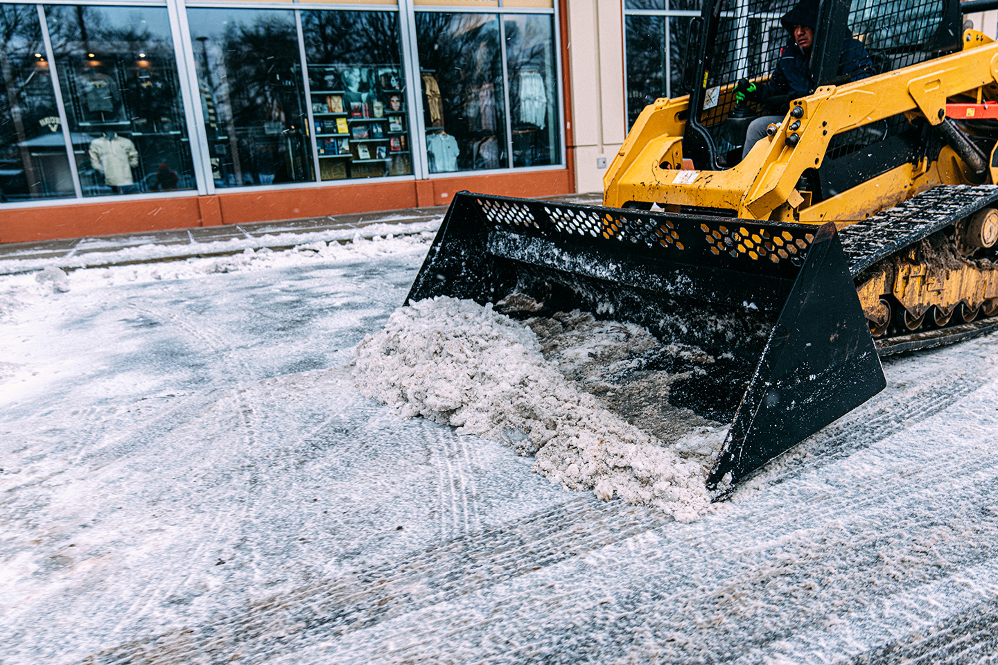 A yellow compact track loader with a black bucket attachment is clearing snow from a paved area in front of a building with large windows reflecting the surroundings. The bucket is full of slushy snow and ice.